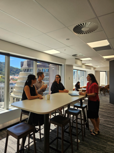 A group of office workers gather around a high table and through the large windows you can see the Beehive.