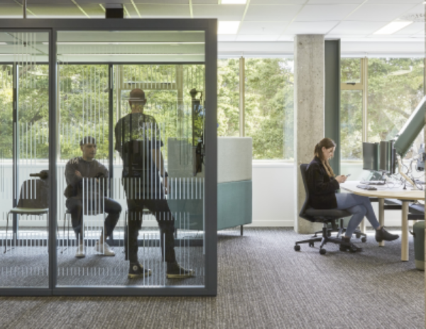 An office space showing two people in a meeting pod and one person sitting at a desk and compputer.